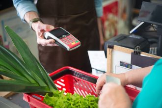 Pessoa realiza pagamento por aproximação em um caixa de mercado, diante de produtos frescos.