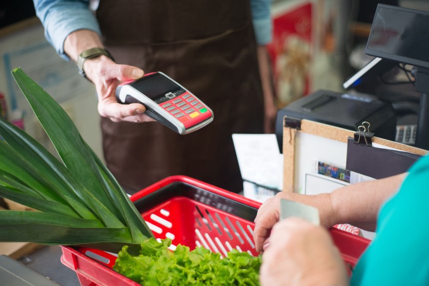 Pessoa realiza pagamento por aproximação em um caixa de mercado, diante de produtos frescos.