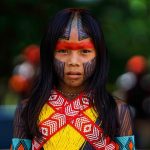 Retrato de uma mulher indígena em ambiente amazônico, com vegetação ao fundo
